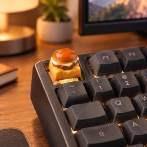 May include: A custom mechanical keyboard keycap shaped like a miniature cheeseburger with a glossy brown bun, a dark brown patty, and a yellow cheese slice, positioned on the top left corner of a black keyboard with backlit keys.
