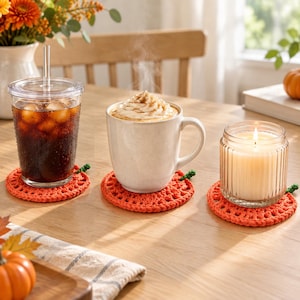 Pode incluir: Tr&ecirc;s bases para copos redondas de croch&ecirc; na cor laranja, em formato de ab&oacute;bora, com pequenos cabos verdes. Uma base sustenta um copo de pl&aacute;stico transparente com caf&eacute; gelado, uma uma caneca de cer&acirc;mica branca e uma uma vela de vidro canelado.
