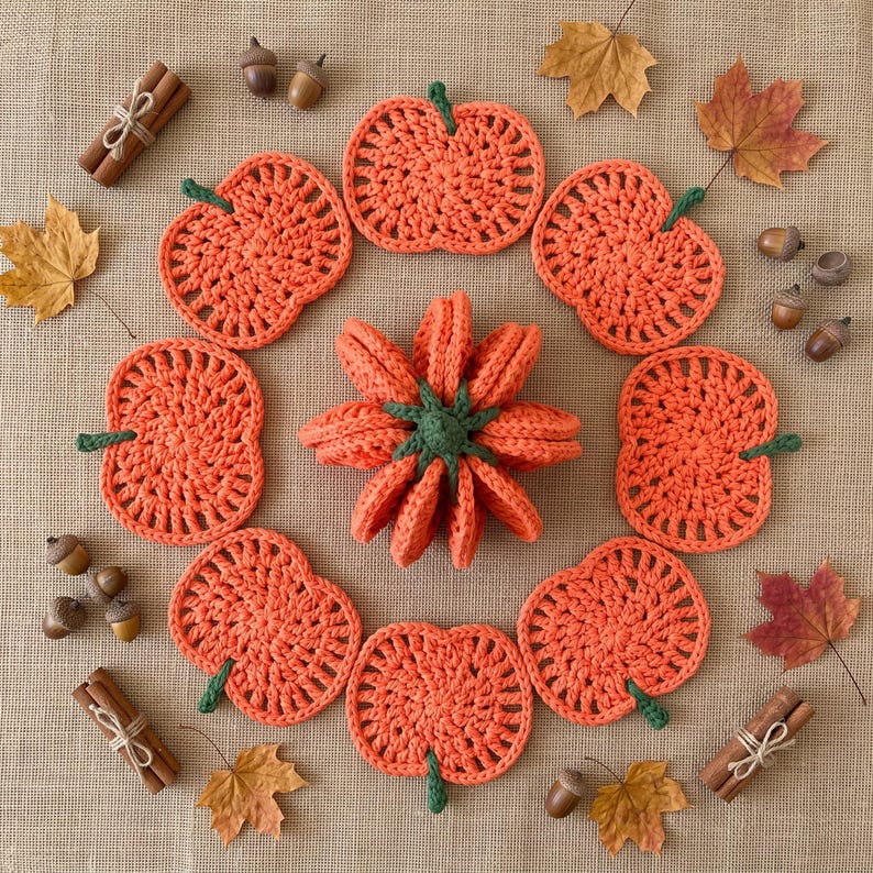 May include: A collection of orange crocheted pumpkin coasters arranged in a circle around a central three-dimensional crocheted pumpkin, accented with scattered brown acorns, cinnamon sticks tied with twine, and dried autumn leaves.