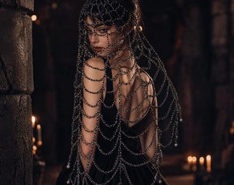 May include: A black chain headpiece featuring a web-like design that drapes over the head and shoulders. The metallic links form a delicate, interconnected pattern, accented with small white beads at the ends of the hanging chain strands.