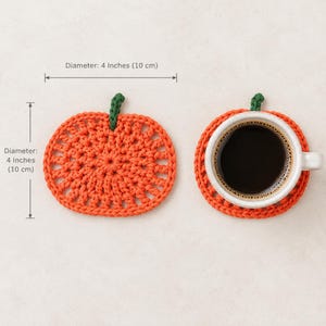 May include: Two orange pumpkin-shaped crochet coasters with green stems. Each coaster measures 4 inches in diameter. One coaster is shown alone, and the other supports a white ceramic mug filled with black coffee, as indicated by the text.