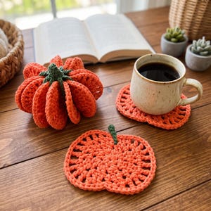May include: A three-dimensional crocheted orange pumpkin with a green stem, alongside two flat, pumpkin-shaped crocheted coasters in the same orange yarn with small green stems, one of which supports a speckled ceramic mug filled with black coffee.