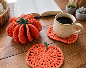 May include: A three-dimensional crocheted orange pumpkin with a green stem, alongside two flat, pumpkin-shaped crocheted coasters in the same orange yarn with small green stems, one of which supports a speckled ceramic mug filled with black coffee.