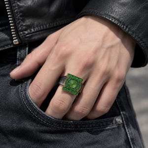 May include: A silver-toned metal ring featuring a rectangular face with a central circular emblem. The emblem and the rectangular border are inlaid with small, vibrant green stones, while the center displays a circular green pattern.