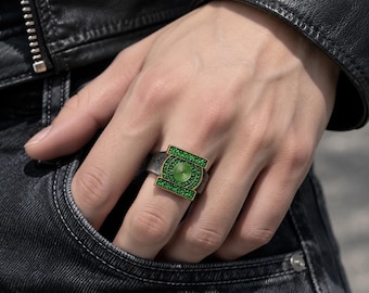 May include: A silver-toned metal ring featuring a rectangular face with a central circular emblem. The emblem and the rectangular border are inlaid with small, vibrant green stones, while the center displays a circular green pattern.