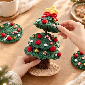 May include: A tiered crochet Christmas tree with a brown trunk and beige base, decorated with red pom-poms, gold bells, white snowflakes, and miniature candy canes. A yellow star sits on top, and the tree is accompanied by matching round coasters.