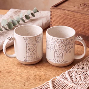 May include: Two off-white speckled ceramic mugs with a knot-shaped handle. One mug features a tree trunk design, the other a full tree with heart-shaped leaves. Both mugs are engraved with a heart containing the letters L+P and the date 2.14.25.