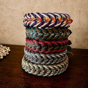 Può includere: Una pila di cinque collari spessi in paracord intrecciato. Il collare superiore presenta motivi viola, bianchi e arcobaleno. Il secondo è verde acqua e marrone. Il terzo è rosso e verde oliva. Il quarto è grigio e nero. Quello in basso è verde menta e marrone.
