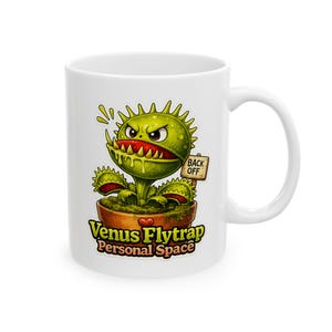 May include: A white ceramic mug featuring a green Venus flytrap illustration in a brown pot. The plant has a red interior, sharp teeth, and a sign that reads BACK OFF. Below the pot, the text reads VENUS FLYTRAP Personal Space in yellow and orange.