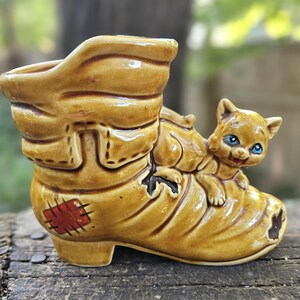 May include: A glazed ceramic planter shaped like a worn boot with a patch on the side and a small cat figure attached to the front. The boot is tan with brown accents, and the cat has blue eyes and a tan coat, creating a decorative vessel.