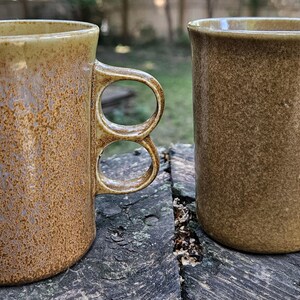 May include: Two ceramic mugs with a speckled brown and tan glaze. Each mug features a unique handle design consisting of two stacked circular loops. The left mug has a lighter, slightly lavender-toned base, while the right mug is a solid earthy brown.
