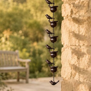 May include: A dark brown metal rain chain featuring a series of connected cups and small bird figurines. The vertical structure hangs from a hook, alternating between open-top bowls and perched bird accents, creating a decorative water feature.