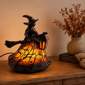 May include: A decorative lamp shaped like a witch wearing a pointed hat and holding a broomstick with a lantern. The base of the lamp features a translucent orange glow with a black silhouette pattern of bare tree branches and spiders.