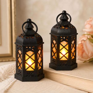 May include: Two black metal lanterns with a hexagonal shape and a lattice pattern on the glass panels. Each lantern features a top ring handle and a decorative cutout design along the roof, containing a warm glowing LED light inside.