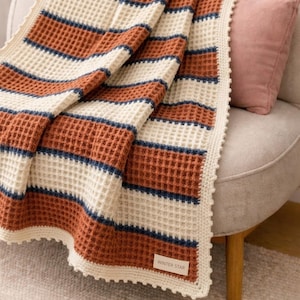 Puede incluir: Una manta de punto tipo gofre con rayas horizontales en color crema, naranja quemado y azul oscuro. La manta tiene un borde de ganchillo color crema y una pequeña etiqueta rectangular en la esquina inferior con el texto WINTER STAR en negro.