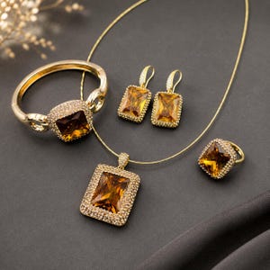 May include: A gold-toned jewelry set consisting of a hinged bracelet, a pair of drop earrings, a pendant necklace, and a ring. Each piece features a large, rectangular, amber-colored faceted gemstone surrounded by a border of small white crystals.