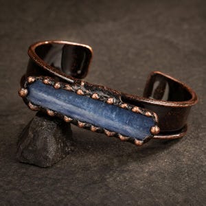 Puede incluir: Una pulsera tipo brazalete con una gema de cianita azul larga y rectangular engastada en un marco de cobre oscuro oxidado. El engaste de metal tiene una apariencia texturizada y rústica con pequeños detalles redondeados en los bordes.
