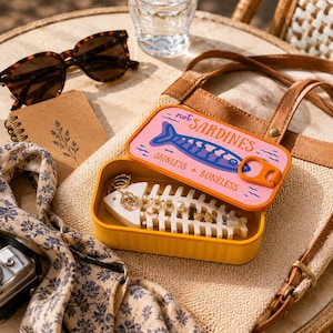 May include: A yellow rectangular jewelry organizer shaped like a sardine tin with a pink lid featuring a blue fish graphic and the text "not SARDINES SKINLESS + BONELESS". Inside, a white fish skeleton-shaped insert holds several rings.