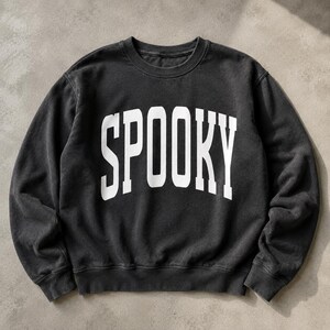 Pode incluir: Uma camisola de gola redonda em cinzento carvão com a palavra SPOOKY impressa em letras maiúsculas grandes, a negrito e brancas no peito, numa fonte universitária arqueada. A peça tem mangas compridas, gola, punhos e bainha canelados.