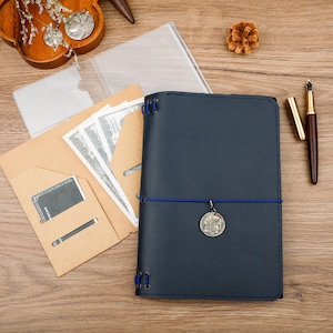 May include: A dark blue leather journal with blue elastic cords and a silver coin charm. It is accompanied by a tan paper insert containing US dollar bills and a black card, a clear plastic sleeve, and a gold-toned fountain pen.