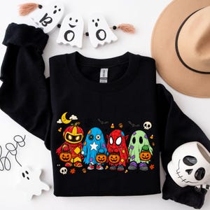Op de afbeelding: Een zwart sweatshirt met lange mouwen en een grafische print van vier figuren in Halloween-kostuums, waaronder een pompoen, een blauwe sterrenheld, een rode spinnenheld en een groene geest, die allemaal oranje pompoenemmers vasthouden.