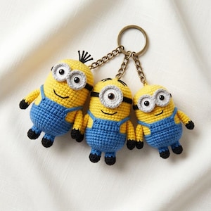 Op de afbeelding: Drie gehaakte Minion-figuren bevestigd aan een enkele bronzen metalen sleutelhanger. Elk figuur heeft een geel lichaam, een blauwe tuinbroek, zwarte handen en voeten, en een wit omrande bril met zwarte ogen. De linkerfiguur heeft zwart haar.