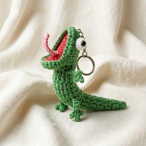 May include: A green crocheted lizard keychain featuring a wide-open mouth with a red interior and a protruding pink tongue. The creature has a single large white eye with a black pupil and is attached to a silver metal split ring key holder.