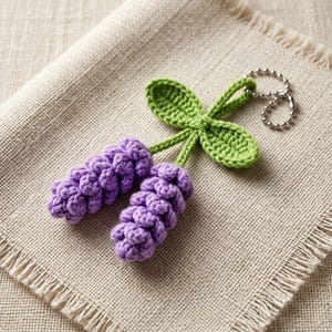 May include: A crochet keychain featuring two textured, light purple lavender flower stalks attached to a pair of bright green crocheted leaves. The floral design is connected to a silver-toned metal ball chain for attachment to keys or bags.
