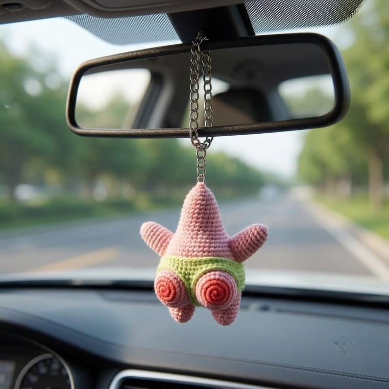 Op de afbeelding: Een gehaakt roze zeester-ornament met een lichtgroene tailleband en twee spiraalvormige uitsteeksels, hangend aan een zilverkleurige metalen ketting bevestigd aan de achteruitkijkspiegel van een voertuig. De figuur heeft een textuur van garen.
