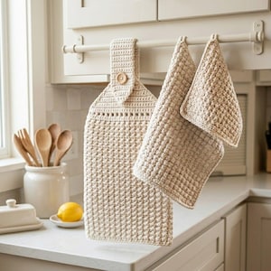 May include: A set of beige crochet kitchen textiles hanging from a rod, including a long towel with a wooden button closure and two smaller square cloths, all featuring a textured, raised stitch pattern in a neutral, light tan yarn color.