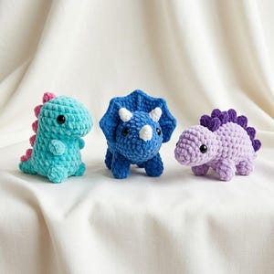 May include: Three crocheted dinosaur plush toys made of soft yarn. The left one is turquoise with pink spikes, the center one is blue with white horns and a frill, and the right one is light purple with dark purple spikes. All have black eyes.