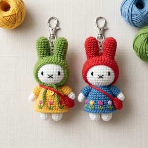 May include: Two crochet rabbit keychains with silver lobster clasps. The left rabbit wears a green hood and yellow dress with embroidered tulips. The right rabbit wears a red hood and blue dress with embroidered tulips. Both have white faces.