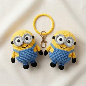 May include: Two yellow and blue crocheted figures resembling characters with large grey goggles, black eyes, and blue overalls. A yellow circular ring with a gold-toned metal lobster clasp is attached to the top of the figures for use as a keychain.