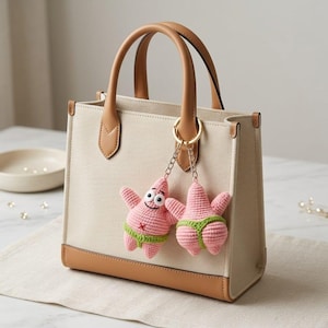Op de afbeelding: Een beige canvas tas met lichtbruine lederen afwerking en twee handvatten. Aan de goudkleurige ring aan het handvat hangen twee roze gehaakte zeester-bedels, die elk een groene string dragen en een lachend gezicht hebben.