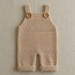 May include: A beige crochet romper featuring a square neckline, two thick shoulder straps, and two round wooden buttons attached at the front. The garment is crafted with a consistent stitch pattern and includes cuffed leg openings at the bottom.