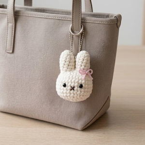 May include: A beige canvas tote bag with a crochet rabbit keychain attached to the handle. The keychain is white with two ears, black bead eyes, a brown stitched cross mouth, and a small light pink bow on the side of its head.