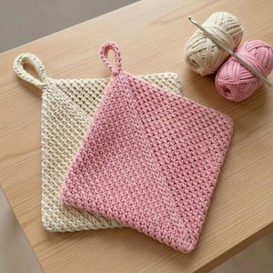 Puede incluir: Dos agarraderas cuadradas de ganchillo, una en color crema y otra en rosa claro, cada una con un lazo para colgar en la esquina. Junto a las agarraderas hay dos ovillos de lana, uno crema y otro rosa, con un ganchillo plateado encima.