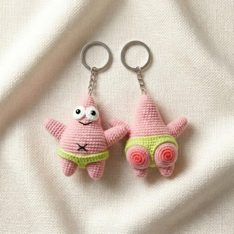 Op de afbeelding: Twee gehaakte sleutelhangers van een roze zeesterfiguur. De ene toont de voorkant met witte ogen en een limoengroene tailleband, de andere toont de achterkant met een limoengroene tailleband en twee roze cirkelvormige details op het onderlichaam.