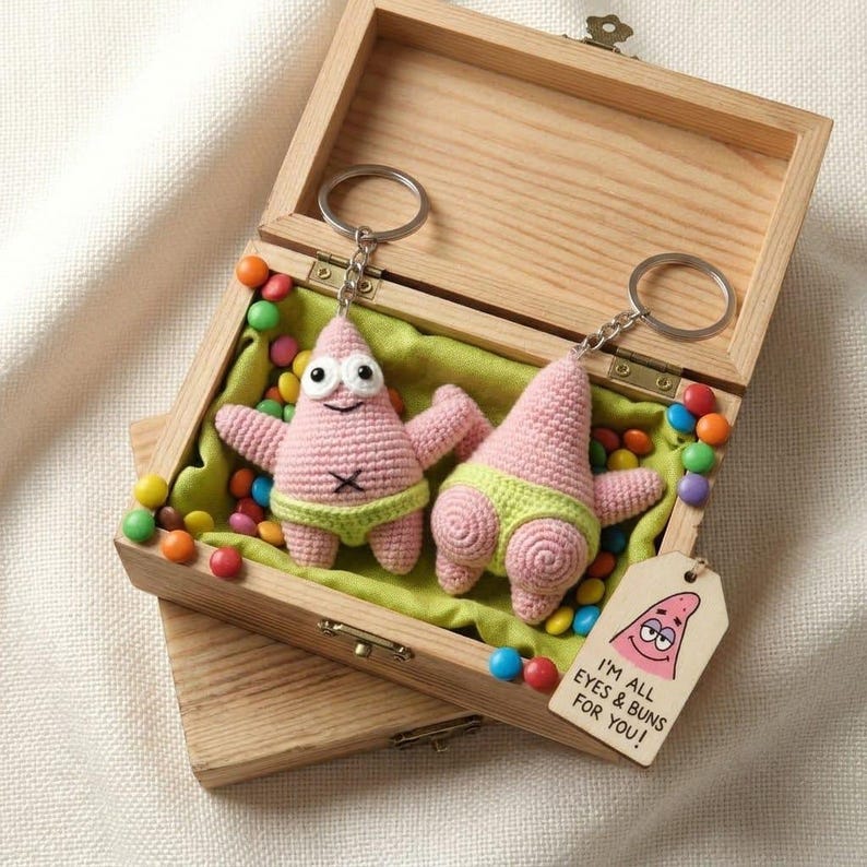 Op de afbeelding: Twee gehaakte roze zeester-sleutelhangers met limoengroene broekjes in een houten doos gevuld met kleurrijke snoepjes. De ene sleutelhanger toont een lachend gezicht, de andere een achterwerk. Een houten label vermeldt: I'M ALL EYES & BUNS FOR YOU!