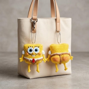 Op de afbeelding: Een beige canvas tas met lichtbruine leren hengsels is voorzien van twee gehaakte gele spons-bedels die aan zilveren kogeltjeskettingen hangen. De ene bedel toont de voorkant, terwijl de andere de achterkant met bruine details laat zien.