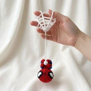 May include: A crocheted ornament featuring a red and blue spherical character with large white eyes, suspended by a long white yarn string from a white crocheted web-like structure. The character has a round red body with blue accents.