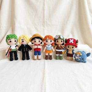 Pode incluir: Um conjunto de seis bonecos de crochê feitos à mão representando vários personagens, incluindo figuras com cabelos verdes, loiros e alaranjados, um chapéu de palha, óculos de proteção e um chapéu vermelho com uma cruz branca, além de um pequeno peluche de crochê em forma de baleia azul.