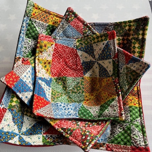 May include: A stack of colorful patchwork quilted items. The items include a square trivet, a smaller square trivet, and two smaller square pot holders. The patchwork features red, blue, green, and yellow fabrics.