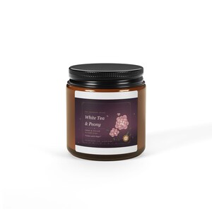 May include: A brown glass candle jar with a black screw-top lid and a dark purple label. The label features white text reading White Tea & Peony, Emma & Julian, and floral illustrations of pink peonies and a small yellow flower on a dark background.