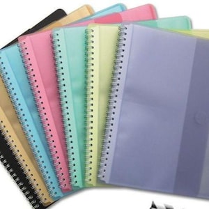 Puede incluir: Una colección de cuadernos de espiral con cubiertas de plástico translúcido en una variedad de colores pastel y sólidos, incluyendo negro, beige, azul, rosa, verde menta, amarillo y morado, dispuestos en forma de abanico superpuesto.