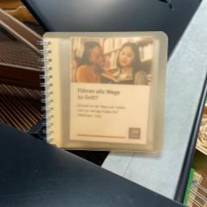Könnte beinhalten: Ein kleines, quadratisches Heft mit Spiralbindung und halbtransparentem Einband. Das Cover zeigt ein Foto von zwei Personen, die in einer Bibliothek lesen, sowie den deutschen Text "Führen alle Wege zu Gott?" und ein kleines Logo.