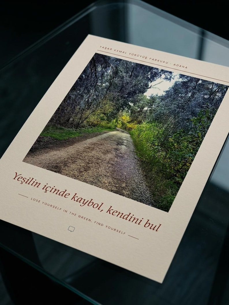 Puede incluir: Una impresi&oacute;n rectangular sobre papel crema con una fotograf&iacute;a de un camino de grava que serpentea a trav&eacute;s de un denso bosque verde. El texto dice Yaşar Kemal Y&uuml;r&uuml;y&uuml;ş Parkuru Adana, Yeşilin i&ccedil;inde kaybol, kendini bul y Lose yourself in the green, find yourself.