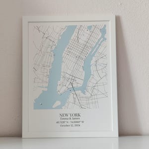 May include: A framed map print of New York City featuring light blue water and black street lines. The text reads NEW YORK, Emma & James, 40.7128&deg; N - 74.0060&deg; W, and October 12, 2024. The print is housed in a simple white frame.