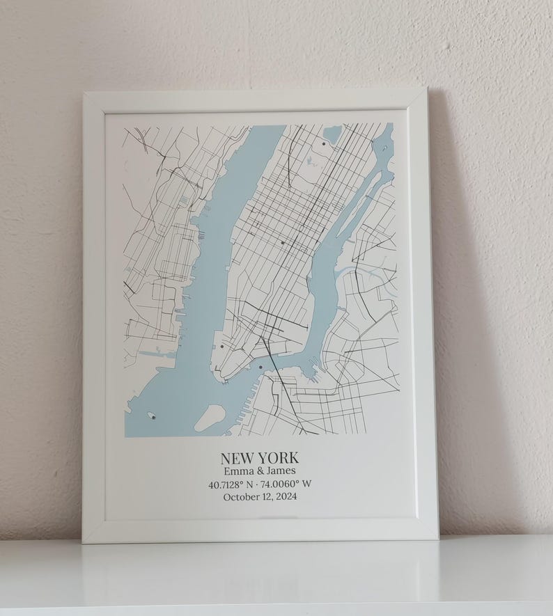 May include: A framed map print of New York City featuring light blue water and black street lines. The text reads NEW YORK, Emma & James, 40.7128&deg; N - 74.0060&deg; W, and October 12, 2024. The print is housed in a simple white frame.