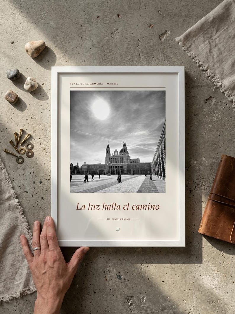 Op de afbeelding: Een ingelijste zwart-wit fotoprint van de Plaza de la Armeria in Madrid. De print bevat de tekst Plaza de la Armeria Madrid bovenaan en La luz halla el camino onderaan, met een klein tekstballon-icoontje onder de tekst.